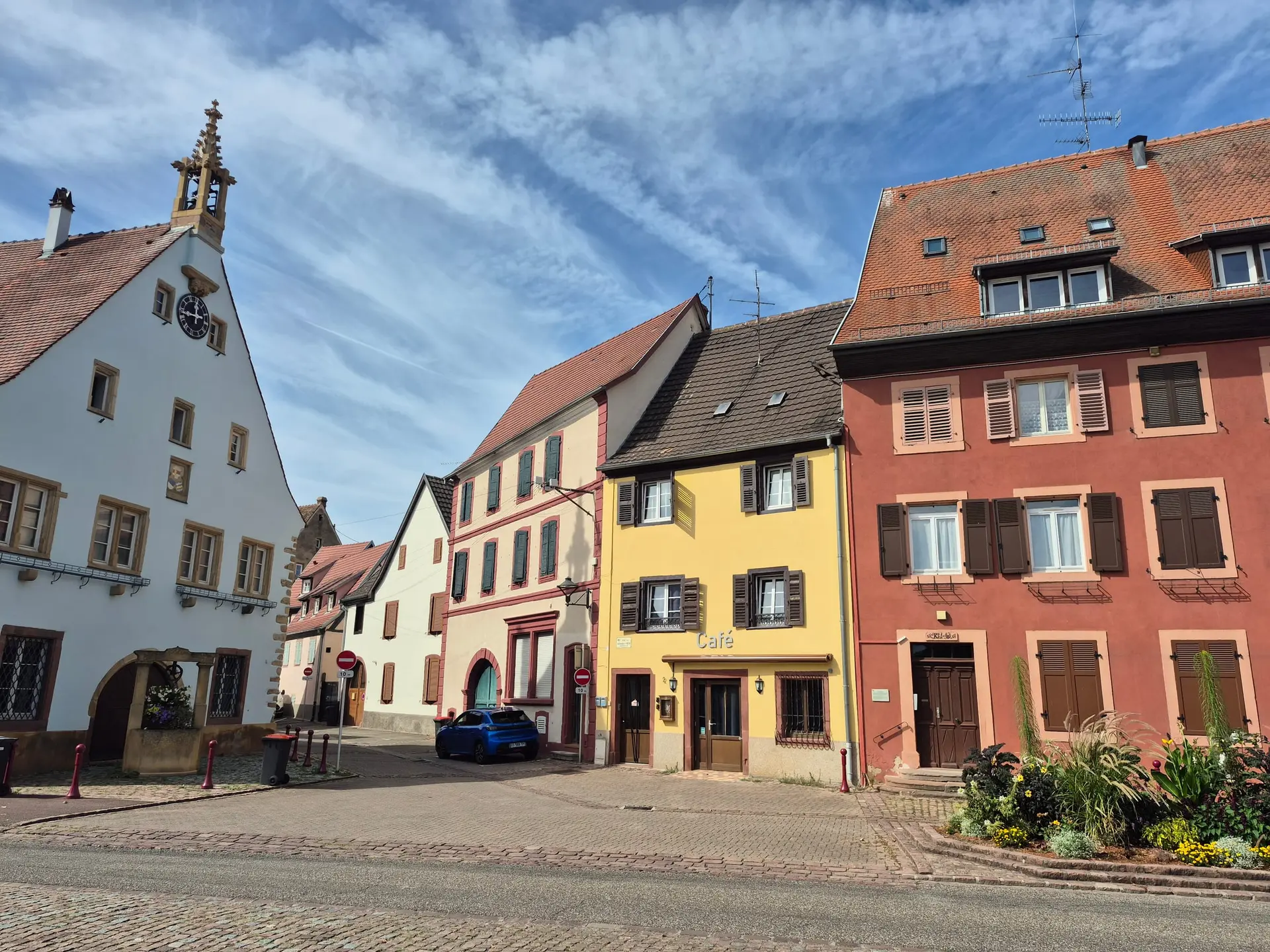 La place du village d'Ingersheim : le bâtiment ancien à horloge et clocheton, et une rangée de maisons alsaciennes jaune, rose et rouge.