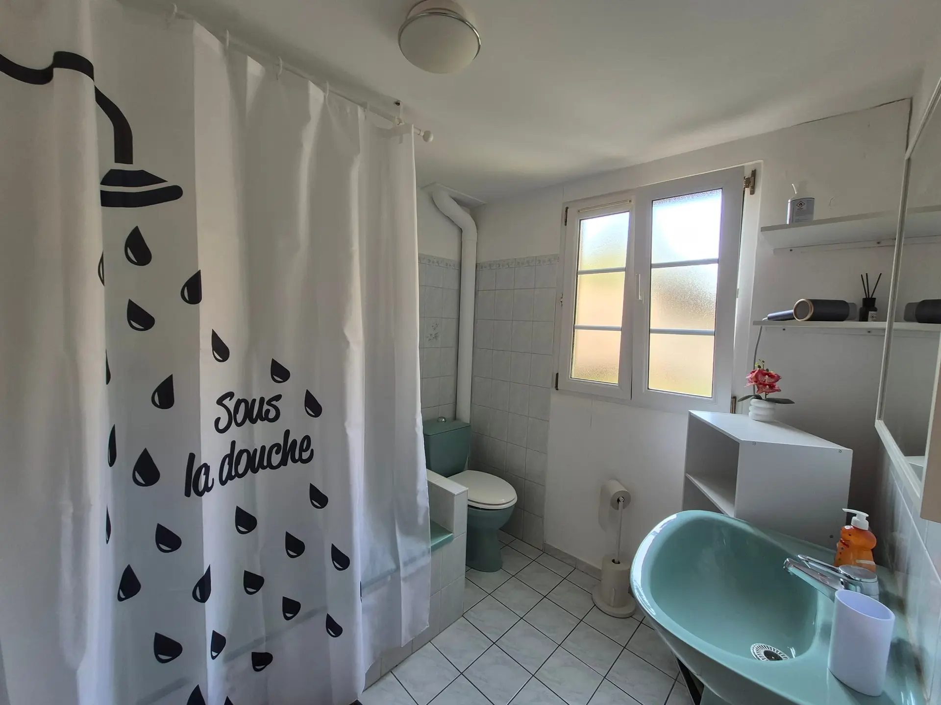 Salle de bain carrelée de blanc, baignoire fermée par un rideau de douche et WC.