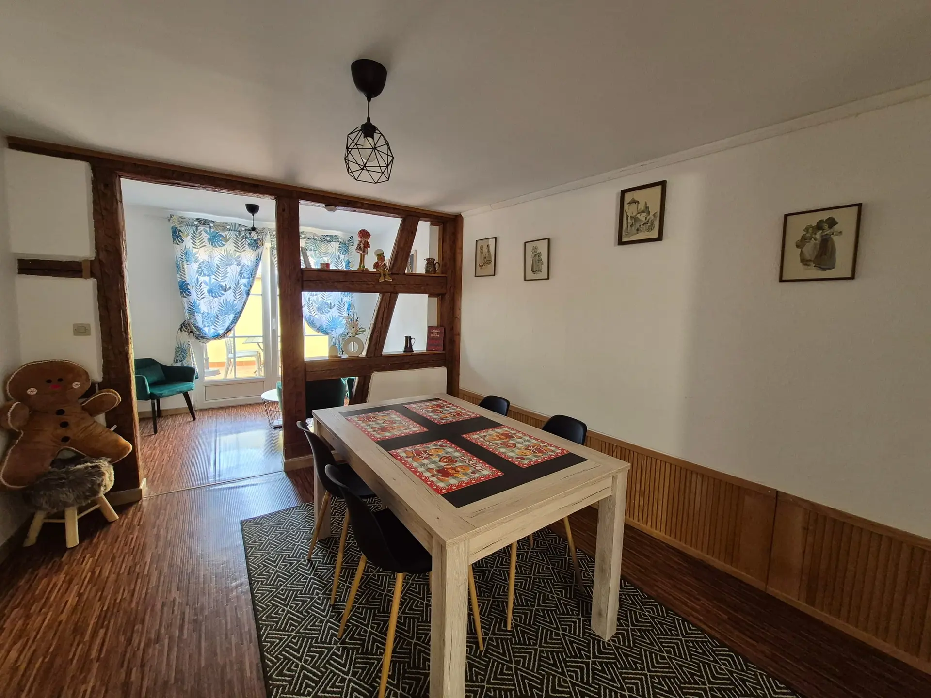 Salon-salle à manger du gîte : table en bois, mur à colombages et fenêtre habillée de rideaux.