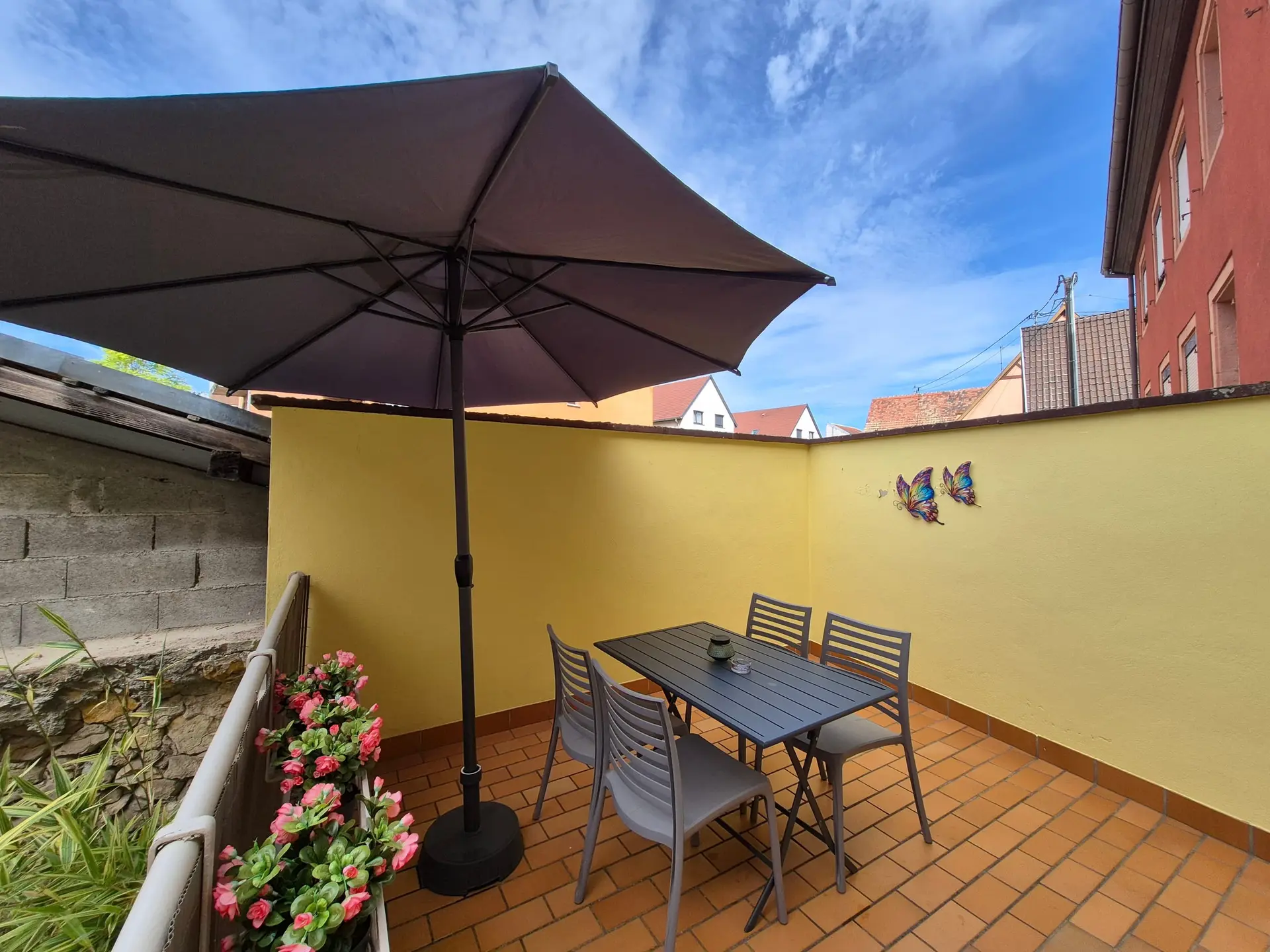 Terrasse carrelée aux murs jaunes, avec table et quatre chaises de jardin.