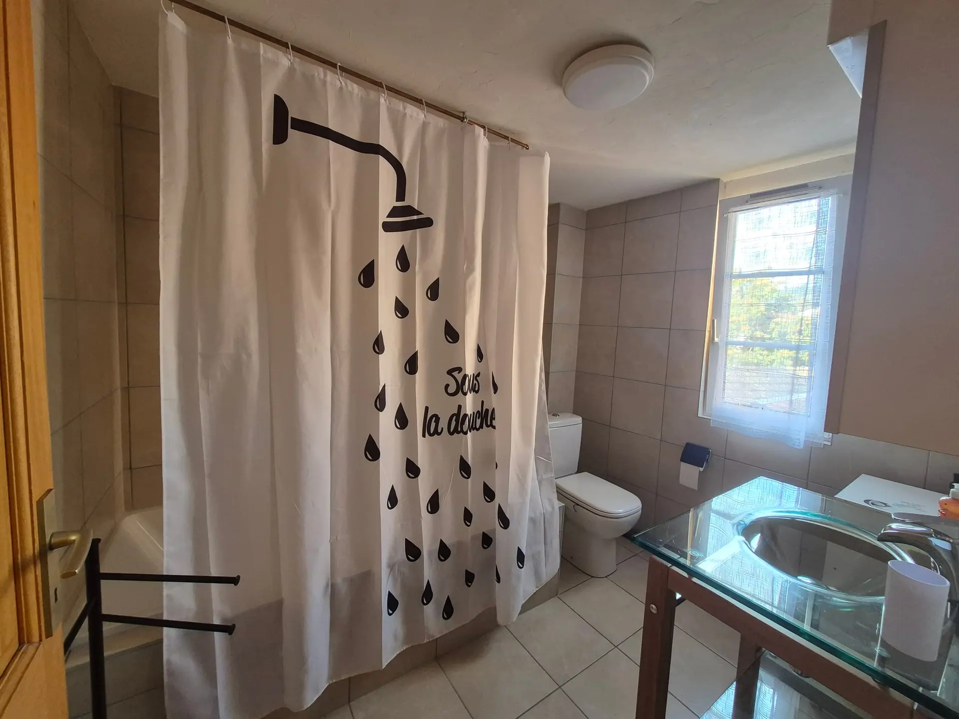 Salle de bain carrelée : baignoire derrière un rideau de douche, WC et lavabo sur un meuble en verre.