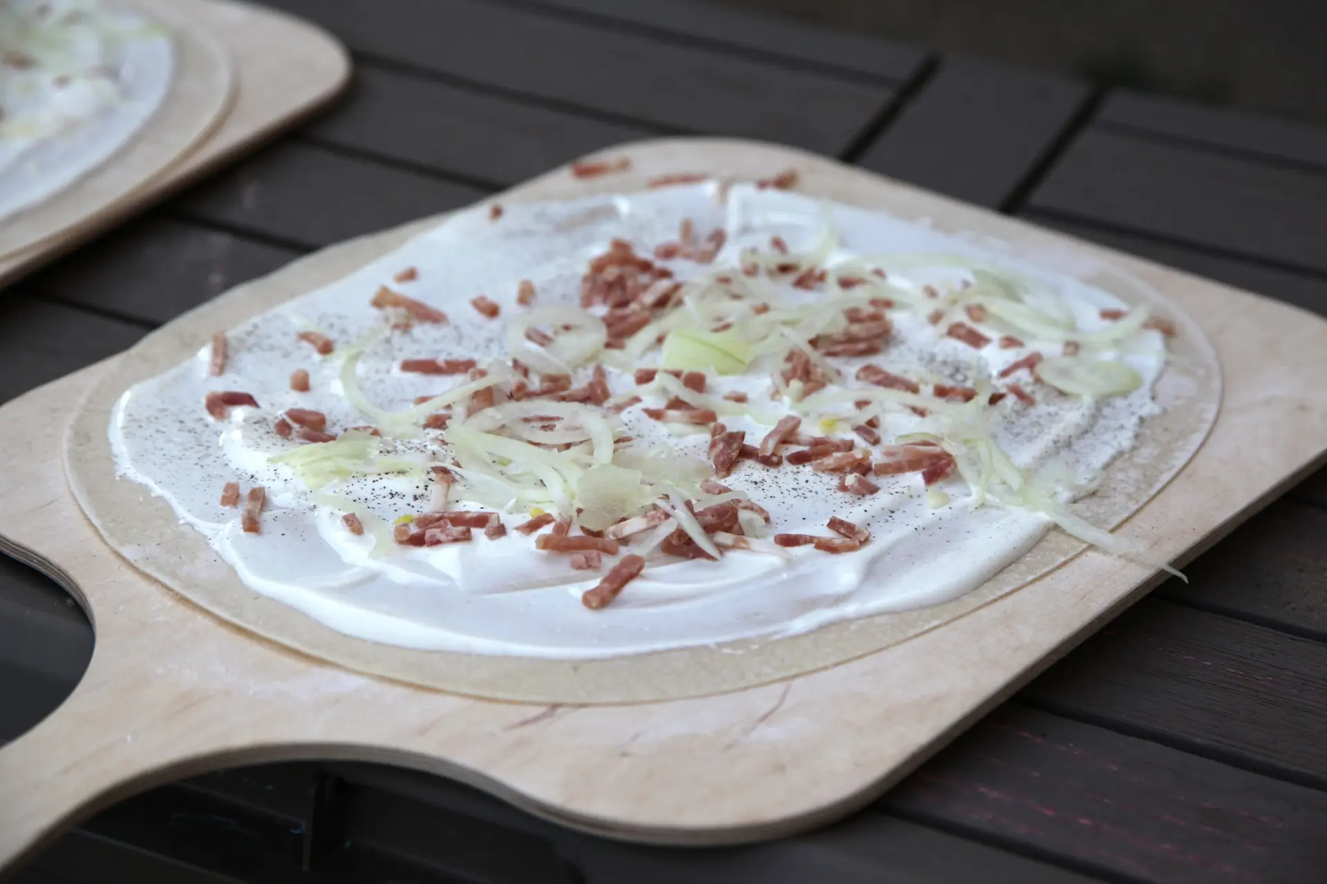 Une tarte flambée garnie de crème, de lardons et d'oignons, prête à enfourner sur sa planche en bois.