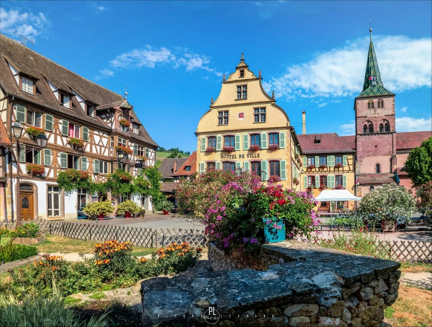 L'hôtel de ville de Turckheim, sa façade jaune à pignon et le clocher de l'église, derrière un jardin fleuri.